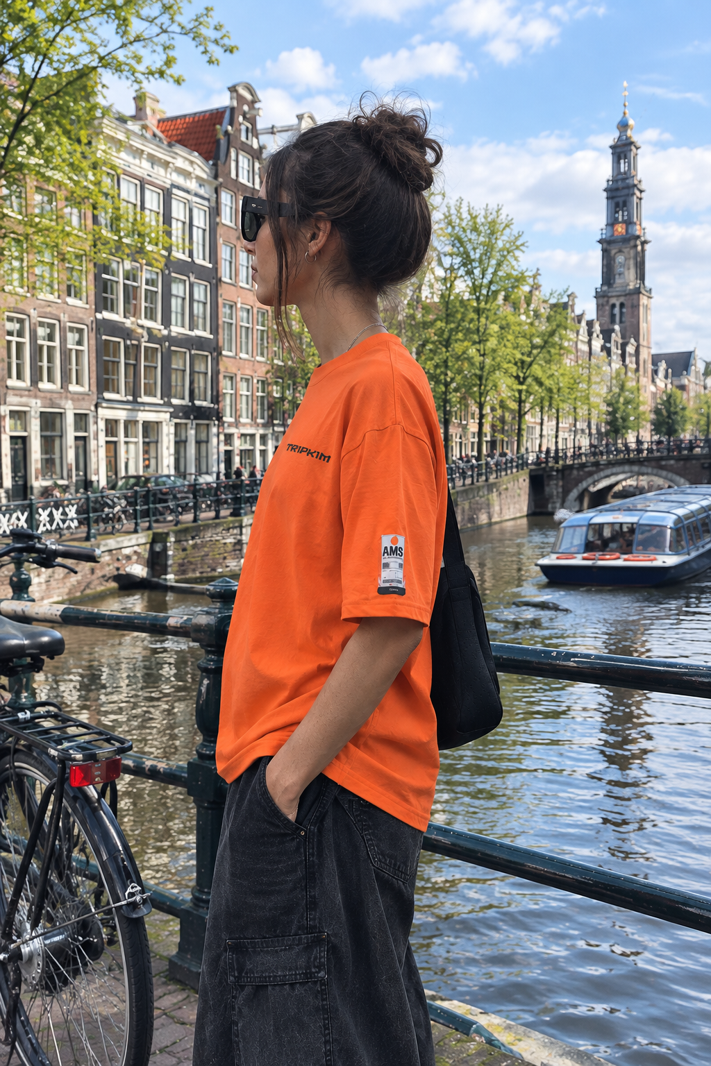 Femme portant le t-shirt Amsterdam AMS orange au bord d'un canal - TRIPKIM